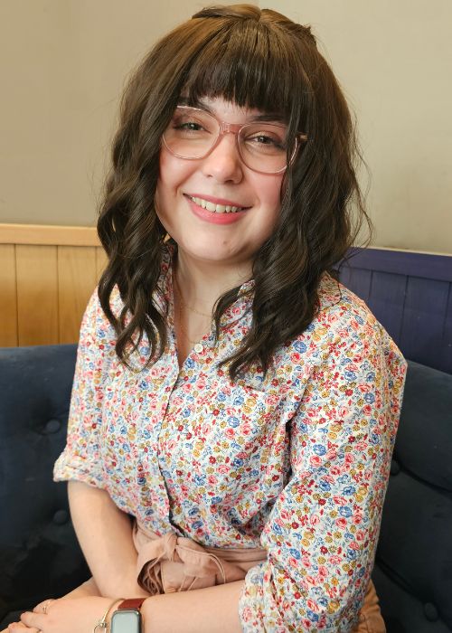 Young woman with brown hair and pink glasses, sat looking at the camera smiling.