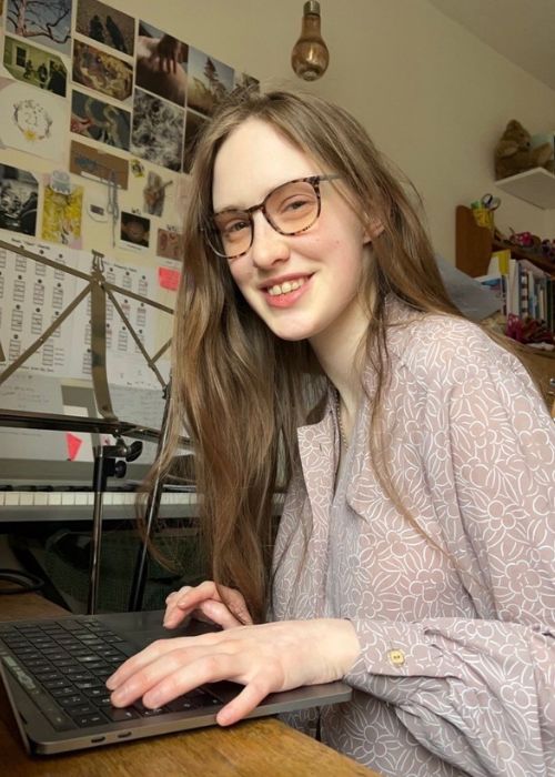 Young woman with brown hair wearing glasses, smiling at the camera as she works on laptop.