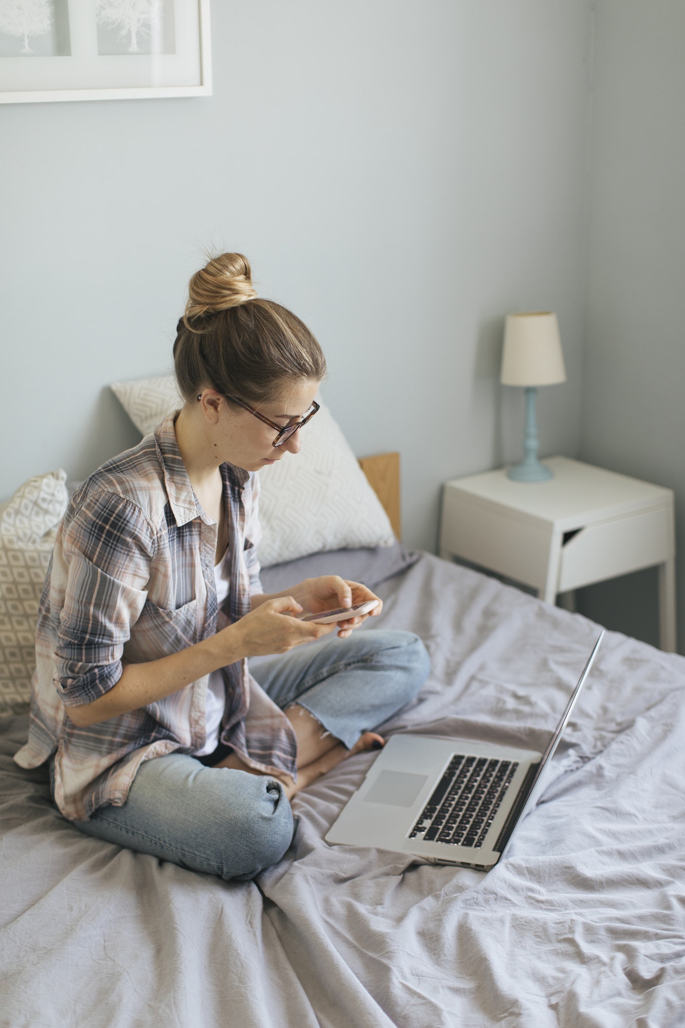 Woman sat with laptop on bed, working from home using phone