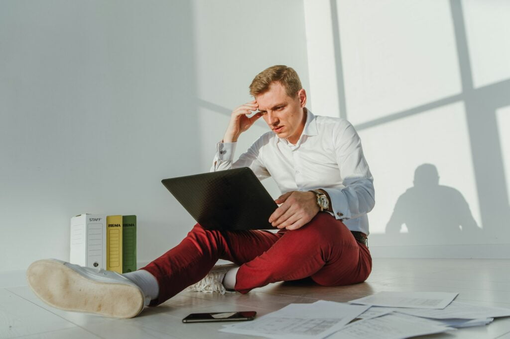 A young man sitting on the floor, surrounded by paper documents and a laptop on his lap. He's looking confused - SEO is made out to be challenging! Our 7 steps to improve your page's SEO simplifies it.