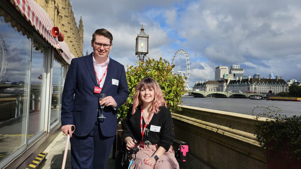 Cameron and Imali at the Small Business Britain event at the House of Lords. It's a sunny day as they're stood by the River Thames. Cameron is wearing a smart white shirt and a blue suit, holding a glass of champagne and leaning on a walking stick. Imali has pink hair and is sitting in her electric wheelchair. She's wearing a pink polkadot dress and a black cardigan.