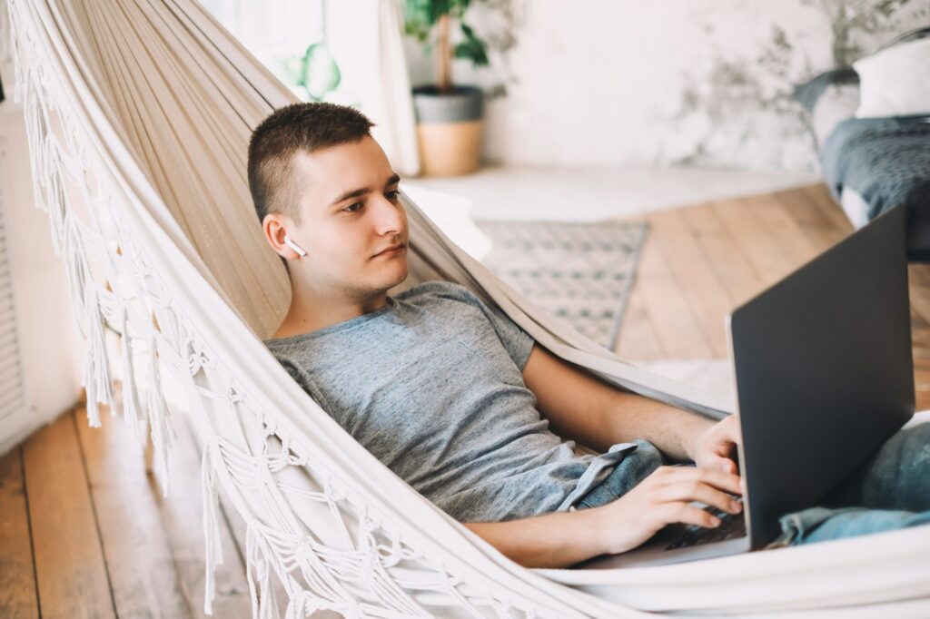 A young man working on his business from a laptop in a hammock. He looks relaxed