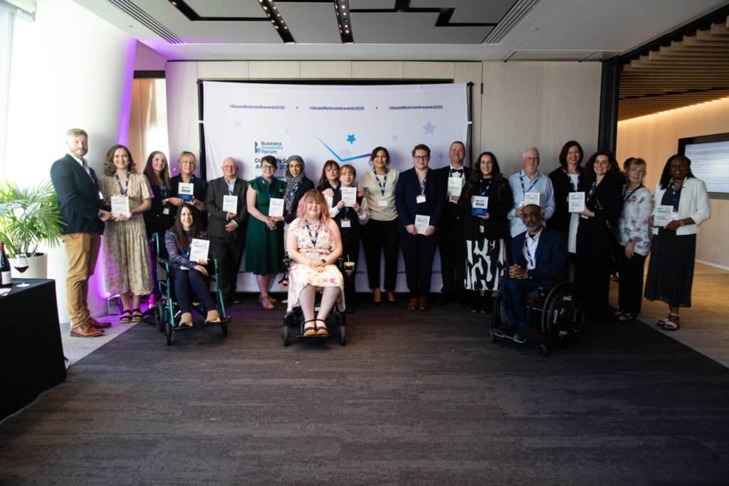 A group photo of the 2025 Disability Smart Award winners standing and seated in front of a branded backdrop displaying the Business Disability Forum logo and the hashtag #DisabilitySmartAwards2025. Twenty-one people are present, holding plaques or smiling proudly. Two individuals are seated in wheelchairs at the front, including a young woman in a floral dress with pink hair and a man in a suit. Another woman is seated in a green wheelchair on the left. The atmosphere is professional and celebratory, with soft purple lighting on the left and natural light coming from the windows.
Inkfire wins Disability Smart Award 2025