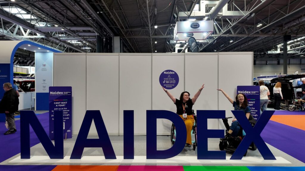A blue text photospot at Naidex, with the words 'Naidex'. Behind the letters sit two wheelchair users, with their hands in the air.